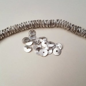 May include: A strand of silver beads, each with a unique, wavy, organic shape, lies on a white surface. A pile of smaller, flat, silver discs with a hole in the center are scattered nearby.