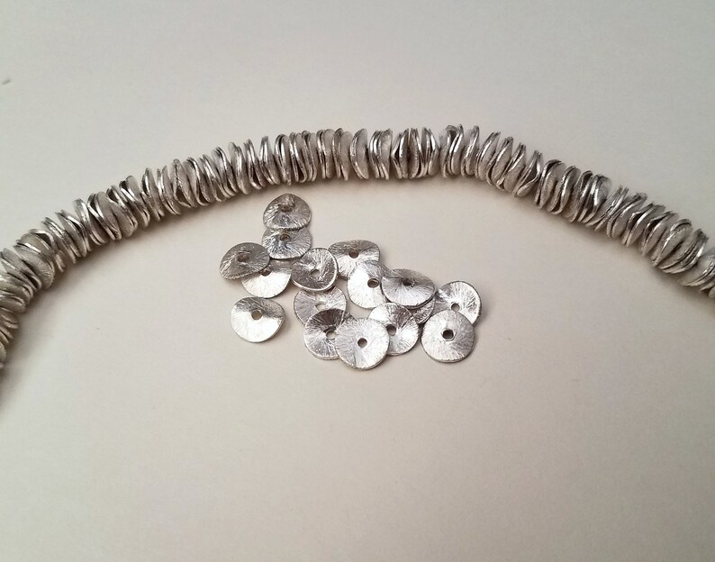 May include: A strand of silver beads, each with a unique, wavy, organic shape, lies on a white surface. A pile of smaller, flat, silver discs with a hole in the center are scattered nearby.