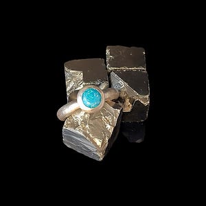 Könnte beinhalten: Ein silberner Ring mit einem runden, hellblauen Stein, der auf einer strukturierten, goldfarbenen Oberfläche gefasst ist. Der Ring wird vor einem dunklen Hintergrund präsentiert, der die Details des Schmucks und die Farbe des Steins hervorhebt.