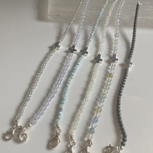 Op de afbeelding: Zes kralenarmbanden met zilveren kruisbedels. De armbanden hebben verschillende gekleurde kralen, waaronder lichtblauw, turquoise, wit en grijs.