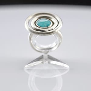 Könnte beinhalten: Ein silberner Ring mit einem türkisfarbenen Edelstein, der in einem konzentrischen Kreisdesign eingefasst ist.