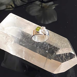 Könnte beinhalten: Ein silberner Ring mit einem runden, hellgrünen Edelstein und zwei kleineren, klaren Edelsteinen. Der Ring liegt auf einem großen, klaren Quarz-Kristall vor dunklem Hintergrund.