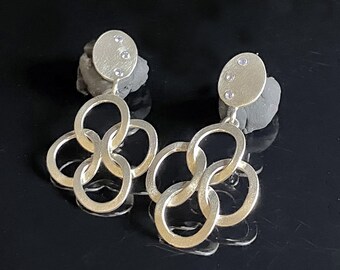 May include: A pair of silver-toned earrings. Each earring features a small, round, textured stud with three small, clear stones. Below the stud, a series of interlocking oval links create a cascading design. The earrings are displayed on a dark surface.