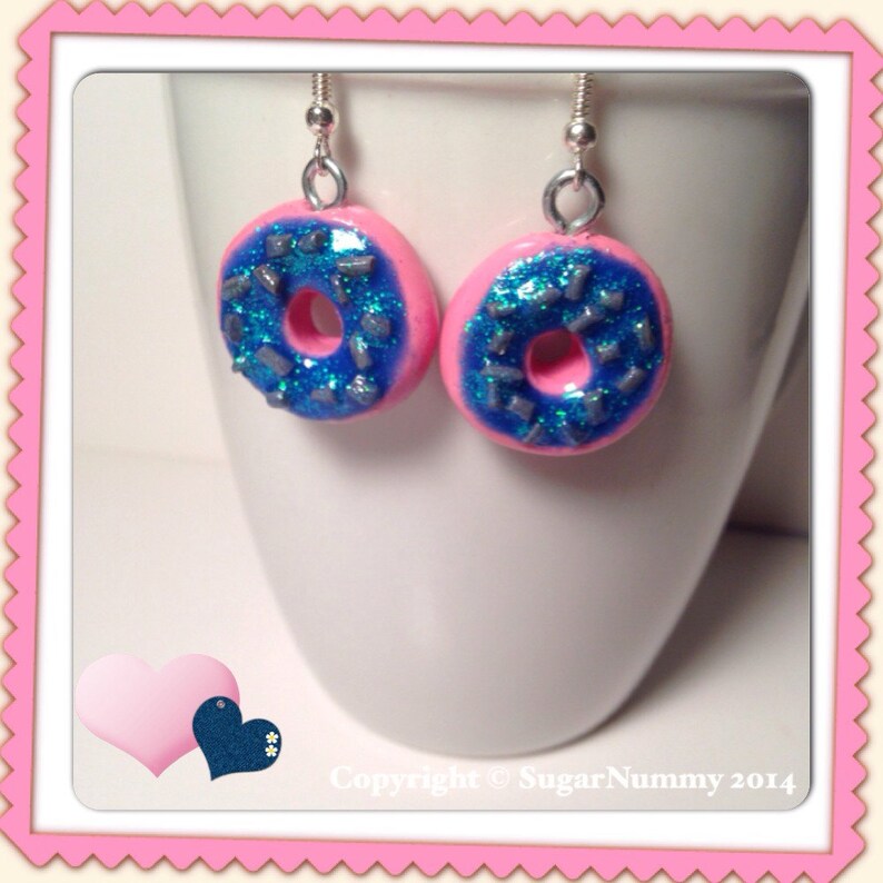 May include: A pair of pink donut earrings with blue frosting and silver sprinkles. The earrings are hanging from silver hooks.