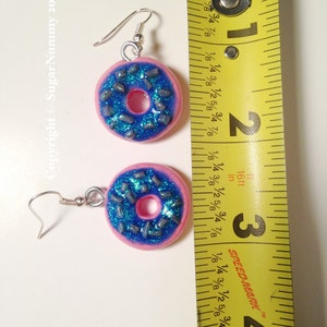 May include: A pair of pink donut earrings with blue frosting and silver sprinkles. The earrings are approximately 1 inch in diameter.
