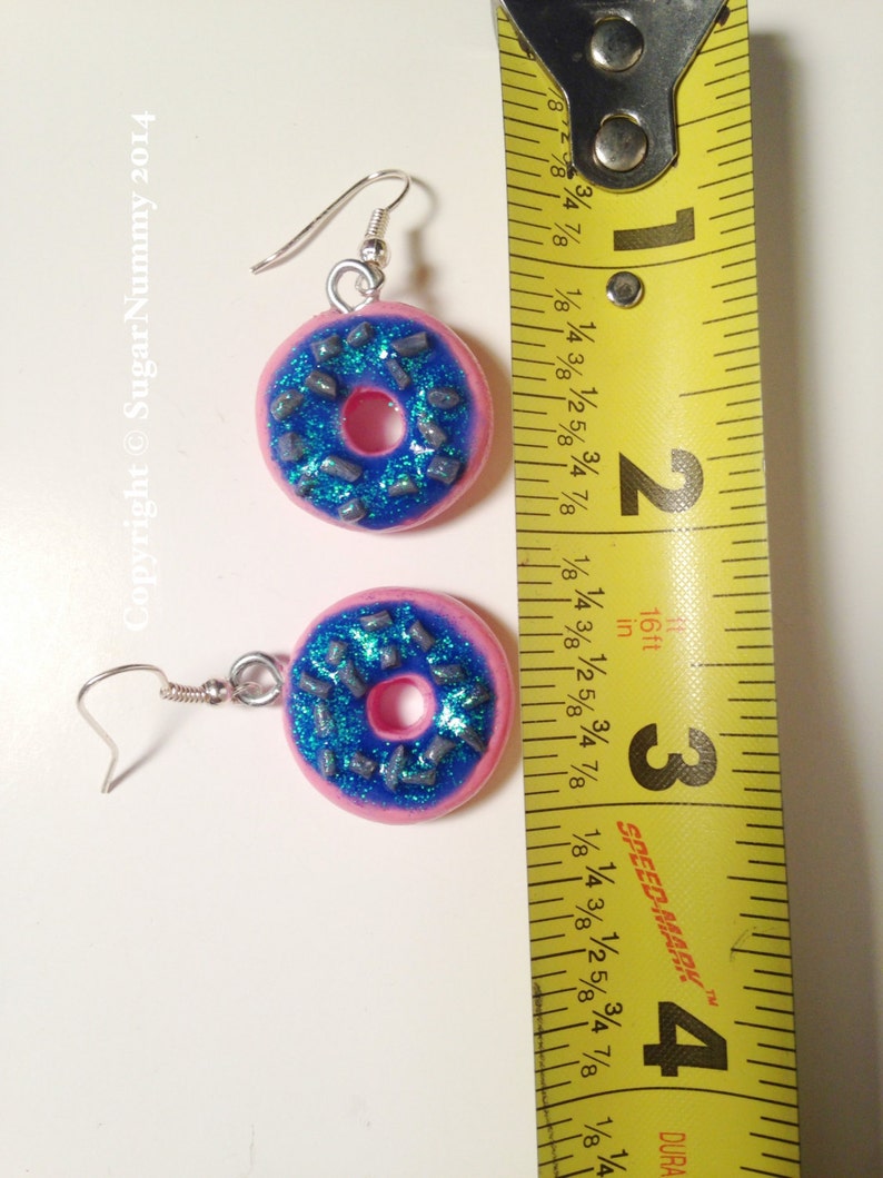 May include: A pair of pink donut earrings with blue frosting and silver sprinkles. The earrings are approximately 1 inch in diameter.
