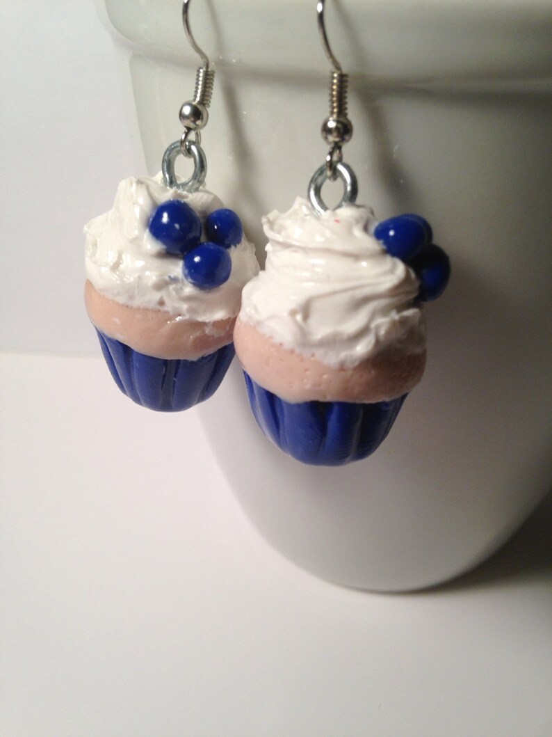 May include: A pair of blue and white cupcake earrings with white frosting and blue blueberry accents.