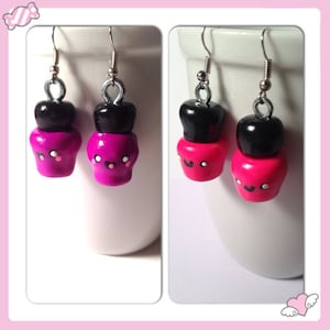 May include: A pair of dangle earrings featuring a black and pink nail polish bottle design. The bottles have a cute cartoon face with white eyes and a pink mouth.