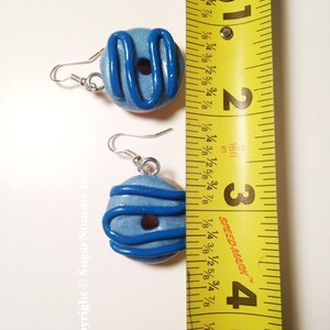 May include: A pair of handmade blue donut earrings with dark blue icing detail. The earrings are shown next to a measuring tape, and are approximately 1 inch in diameter. The earrings have silver-colored ear wires.