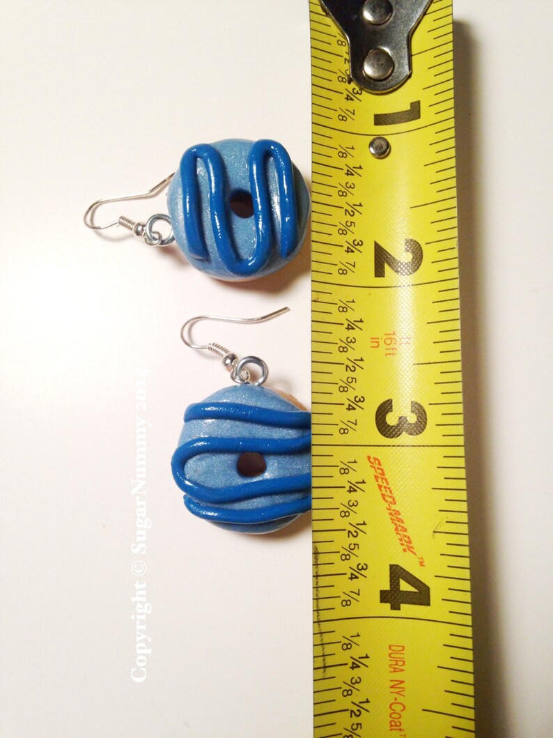 May include: A pair of handmade blue donut earrings with dark blue icing detail. The earrings are shown next to a measuring tape, and are approximately 1 inch in diameter. The earrings have silver-colored ear wires.