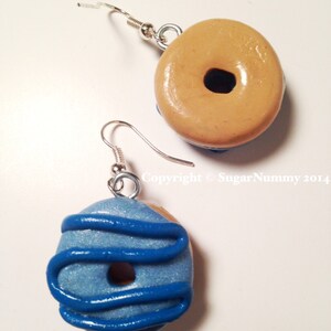 May include: A pair of handmade donut earrings. One earring is tan with a dark brown center, the other is blue with blue icing. Both have silver-colored ear wires. The earrings are a fun, whimsical accessory.
