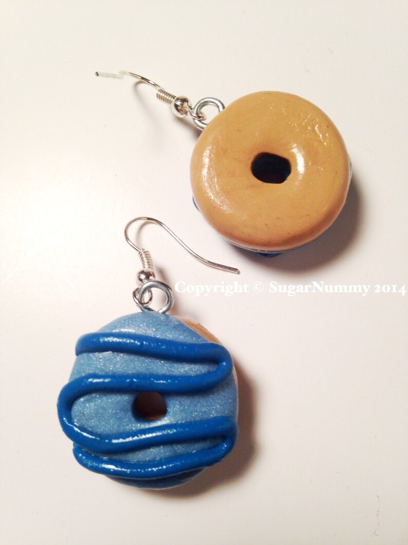 May include: A pair of handmade donut earrings. One earring is tan with a dark brown center, the other is blue with blue icing. Both have silver-colored ear wires. The earrings are a fun, whimsical accessory.