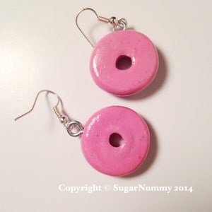 May include: A pair of pink donut earrings with silver hooks. The donuts have a sparkly finish.