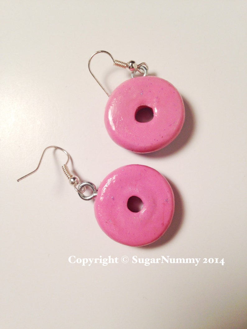 May include: A pair of pink donut earrings with silver hooks. The donuts have a sparkly finish.