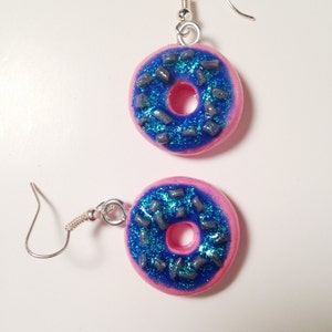 May include: A pair of pink donut earrings with blue frosting and silver sprinkles. The earrings are made of polymer clay and have silver hooks.