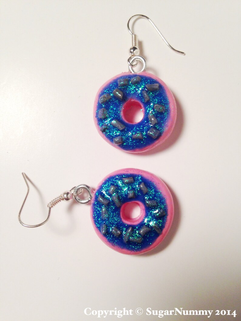 May include: A pair of pink donut earrings with blue frosting and silver sprinkles. The earrings are made of polymer clay and have silver hooks.