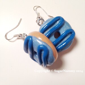 May include: A pair of handmade donut earrings. Each earring features a light blue donut with dark blue icing and a tan-colored donut. The earrings have silver-colored ear wires.