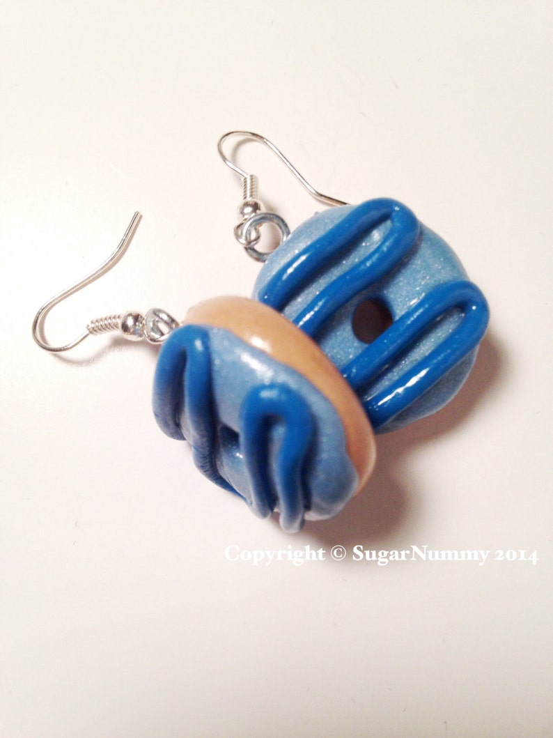 May include: A pair of handmade donut earrings. Each earring features a light blue donut with dark blue icing and a tan-colored donut. The earrings have silver-colored ear wires.