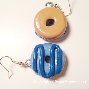 May include: A pair of handmade donut earrings. One earring is tan with a blue base, the other is light blue with blue icing. Both have silver-colored ear wires. The text "Copyright &copy; SugarNummy 2014" is visible.