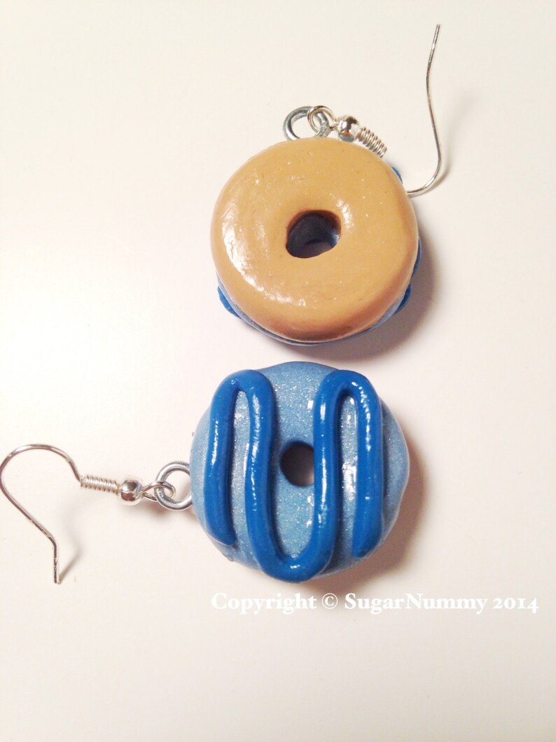 May include: A pair of handmade donut earrings. One earring is tan with a blue base, the other is light blue with blue icing. Both have silver-colored ear wires. The text "Copyright &copy; SugarNummy 2014" is visible.