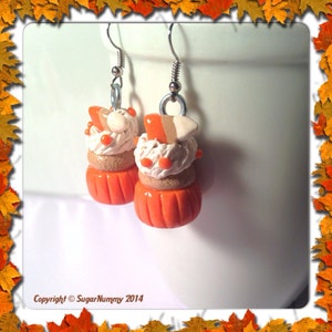 May include: A pair of orange and white pumpkin spice cupcake earrings with silver hooks. The cupcakes are topped with white frosting and a brown and orange candy topping.