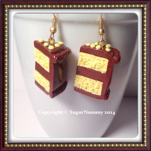 May include: A pair of dangle earrings shaped like a slice of chocolate cake with yellow frosting and sprinkles. The earrings are made of polymer clay and have gold hooks.