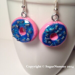 May include: Pink donut shaped earrings with blue frosting and silver sprinkles. The earrings are made of polymer clay and have silver hooks.