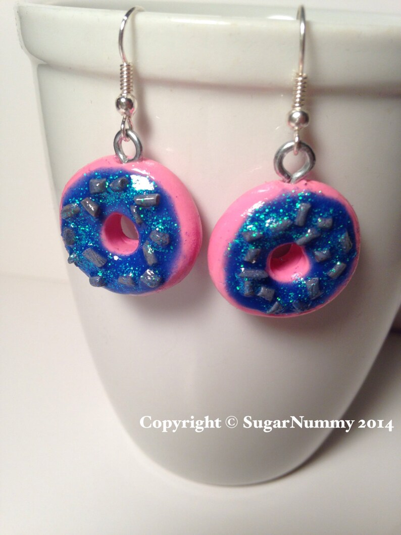 May include: Pink donut shaped earrings with blue frosting and silver sprinkles. The earrings are made of polymer clay and have silver hooks.