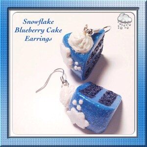 May include: A pair of blue and white snowflake blueberry cake earrings. The earrings are made of polymer clay and are shaped like a slice of cake with white frosting and blue sprinkles. The earrings are attached to silver hooks.