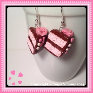 May include: A pair of dangle earrings featuring a chocolate cake slice with pink frosting and a pink rose on top. The cake slices are decorated with white and pink sprinkles.
