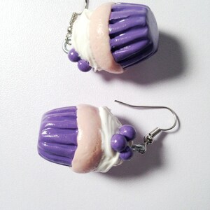 May include: A pair of purple and white cupcake earrings with silver hooks. The cupcakes have a pink layer, white frosting, and purple sprinkles.