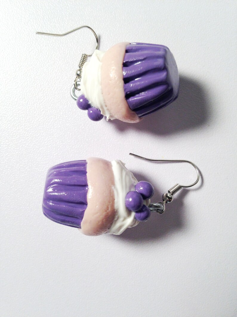 May include: A pair of purple and white cupcake earrings with silver hooks. The cupcakes have a pink layer, white frosting, and purple sprinkles.