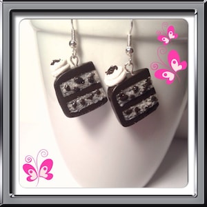 May include: A pair of dangle earrings featuring a chocolate cake slice with white frosting and chocolate chips. The earrings are made of polymer clay and have silver hooks.