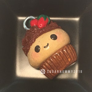 Puede incluir: Un pequeño amuleto de cupcake de arcilla hecho a mano con glaseado de chocolate marrón, una fresa roja y verde en la parte superior y una carita feliz. El amuleto tiene un lazo plateado para colgar.