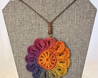 May include: A crochet flower pendant necklace featuring a gradient of colors ranging from deep purple and blue to green, yellow, orange, and red. The flower is attached to a thin brown cord with a knotted detail at the top center.