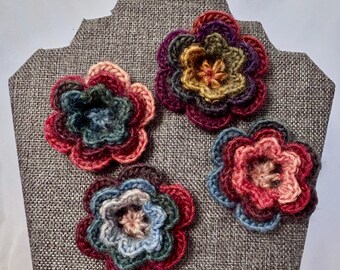May include: Four handcrafted crochet flowers featuring layered petals in variegated yarn colors including deep red, forest green, mustard yellow, soft pink, and slate blue. Each flower has a circular, multi-petaled design with a textured yarn finish.