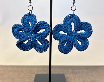 May include: A pair of vibrant blue crochet flower earrings featuring five rounded petals each. The floral motifs are crafted from blue thread and are suspended from dark metal fishhook style earring wires, creating a lightweight accessory.