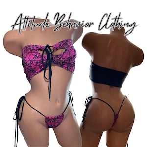 May include: A two-piece bikini set with a pink and black lace pattern. The top is a strapless bandeau style with a lace-up front. The bottom is a thong style with tie sides. The text "Attitude Behavior Clothing" is above the bikini.