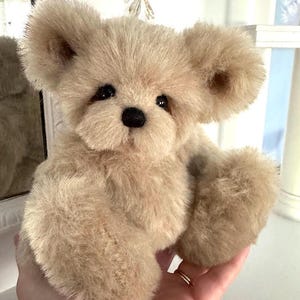 Könnte beinhalten: Ein kuscheliger, hellbrauner Teddybär mit schwarzer Nase und Augen. Der Bär hat große, abgerundete Ohren und eine weiche, pelzige Textur. Er wird in einer Hand gehalten, um seine Größe und Details zu zeigen. Ein klassisches, kuscheliges Spielzeug.