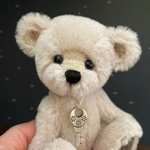 May include: A small, cream-colored teddy bear with a black nose and shiny black eyes. The bear has a silver key pendant around its neck. The bear's paws have brown pads. The bear is a soft, plush toy.
