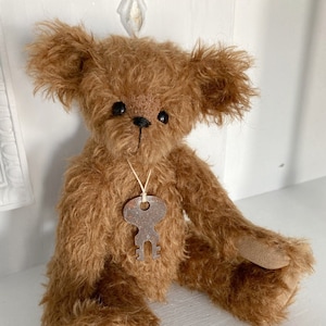 May include: A brown teddy bear with a key charm around its neck. The bear has a fluffy, shaggy coat and is sitting on a white surface.