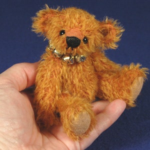 Könnte beinhalten: Ein kleiner, orange-brauner Teddybär mit flauschigem Fell und einer goldenen Glockenhalskette. Der Bär sitzt in der Handfläche.