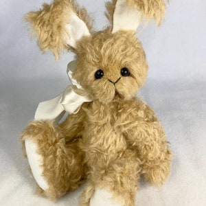 May include: A plush, tan-colored bunny with long, floppy ears and a cream-colored bow. The bunny has a fluffy, textured appearance, with black button eyes and white fabric on the inner ears and feet. It is a soft toy.