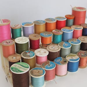 May include: A collection of vintage thread spools in various colors, including pink, green, orange, and blue. The spools are arranged on a wooden display, showcasing the variety of colors and textures. The spools are made of wood and paper.