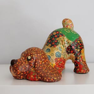 May include: A decorative dog figurine in a playful pose, crafted with a patchwork design. The dog's head and body are adorned with floral patterns in shades of brown, red, green, and yellow. The figurine is placed on a white surface.