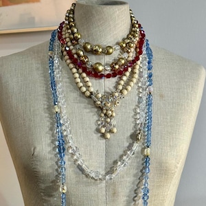 Op de afbeelding: Een verzameling vintage kettingen tentoongesteld op een mannequin. De kettingen hebben verschillende ontwerpen, waaronder strengen rode, witte en gouden kralen, en lange strengen blauwe en heldere kristallen kralen. De kettingen zijn gelaagd.