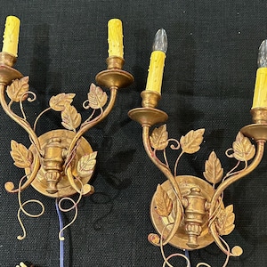 May include: Two antique gold-colored wall sconces with leaf and vine detailing. Each sconce holds two candle-shaped light bulbs. The sconces have a round base and are wired with a blue cord and brown plug.