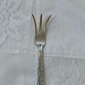 May include: A silver fork with four prongs, set on a white lace tablecloth. The fork's handle features an ornate, decorative design. The prongs are curved and pointed. The overall style is vintage and elegant.
