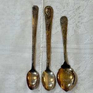 Könnte beinhalten: Drei silberfarbene Vintage-Löffel aus Metall mit angelaufenen Oberflächen. Der linke Löffel hat einen schlanken Griff mit geometrischen Details. Der mittlere Löffel zeigt ein Reliefporträt einer Person. Der rechte Löffel hat eine breite Laffe und ein Muschelmotiv.