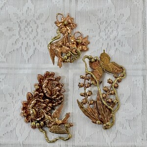 May include: Three copper-toned metal pendants with floral and bird designs. One pendant features a bird with a flower, another has a butterfly, and the third has a flower and bird. The pendants are on a white lace background.